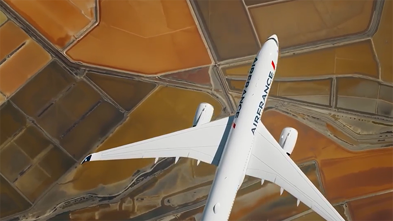An Air France airplane viewed from above flies over a patchwork of green and brown geometric fields. The "AIRFRANCE" logo is visible. 