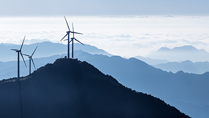 Wind turbines on a mountain ridge set against layered blue mountain ranges and a cloudy sky, symbolizing renewable energy infrastructure and sustainable power solutions.
