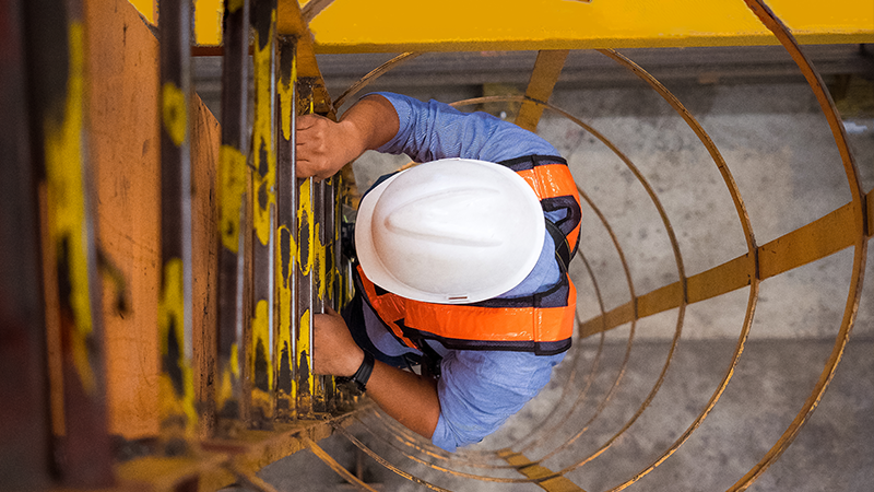 A person in a white hard hat, blue shirt, and orange safety vest climbs a yellow metal ladder with safety cage enclosures, symbolizing real assets and capital solutions in energy infrastructure.